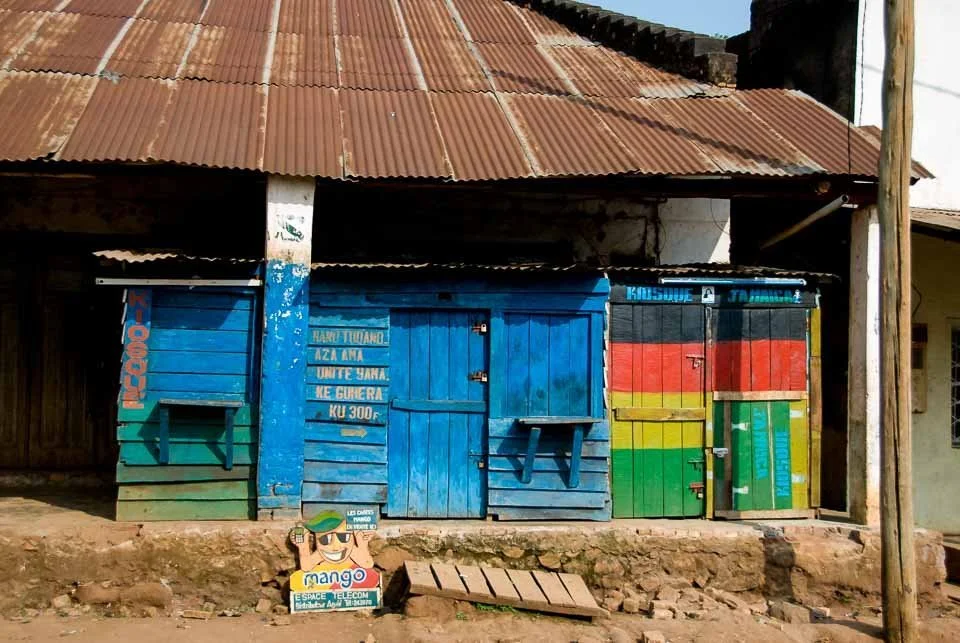 Colorful wooden shops painted in blue, red, yellow, and green in a rural area with a corrugated metal roof, dirt ground, and a sign with a cartoon character advertising a telecom company on the ground.