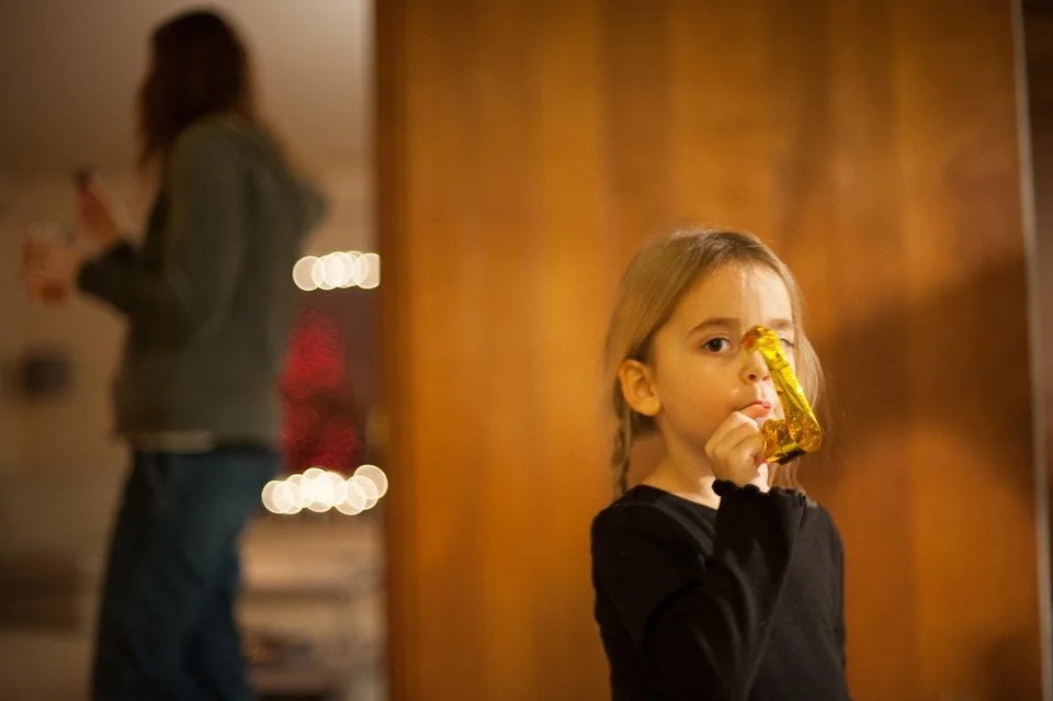 A young girl with long blonde hair, wearing a black shirt, holds a gold party horn to her mouth and looks at the camera. In the background, blurred, is a woman in a gray hoodie holding a drink.