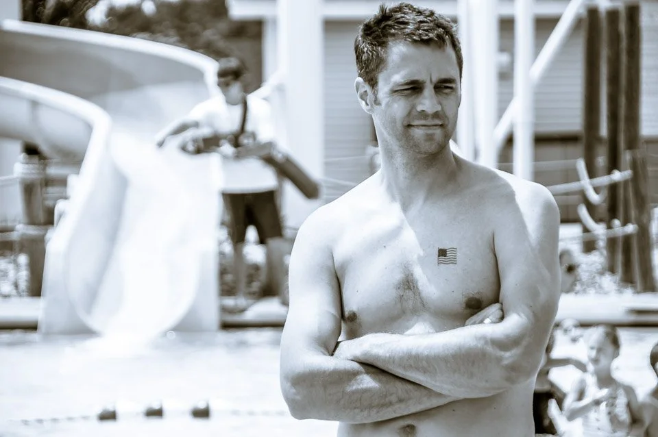 A shirtless man with a small American flag tattoo on his chest, standing outdoors with his arms crossed, looking to the side. In the background, there are children near a pool and a building.