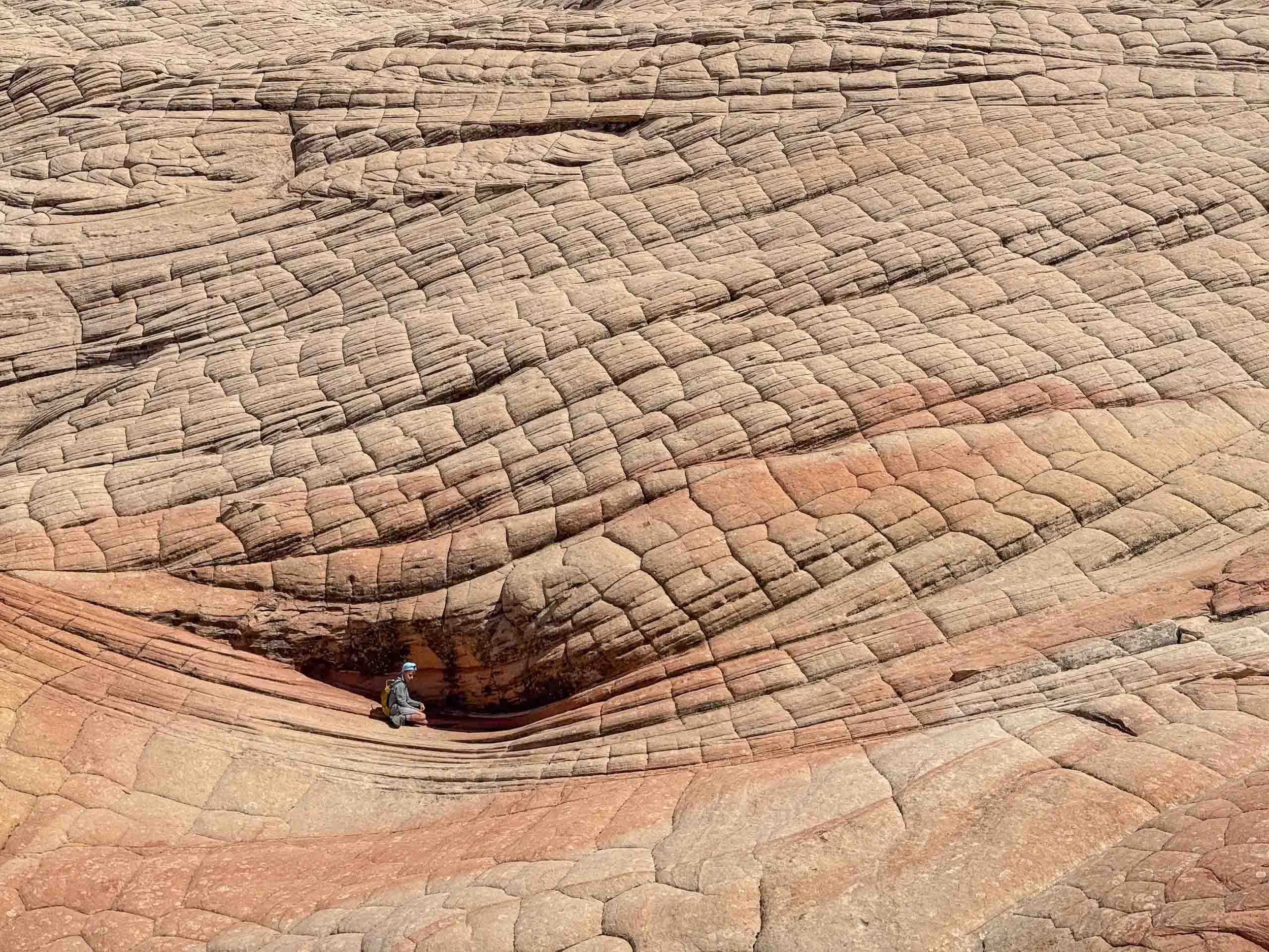 A person in outdoor gear sitting in a small depression on a multicolored rock formation with layered, curved patterns.