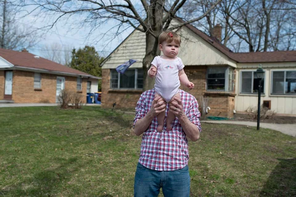 A person holding a baby girl dressed in pink, standing outdoors in a backyard with a house, trees, and a lamp post in the background.