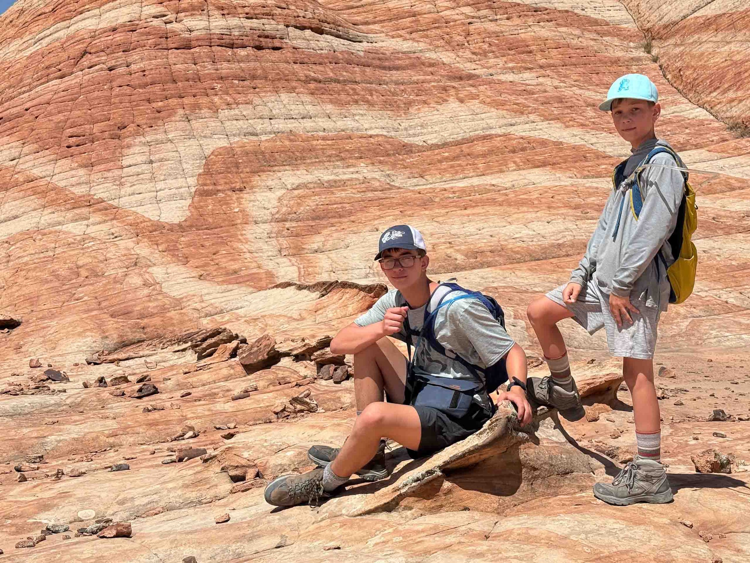 Two boys hiking in a desert with reddish, sandstone cliffs in the background. One boy is sitting on a rock, wearing glasses and a gray cap, while the other stands with one foot on a rock, both carrying backpacks.