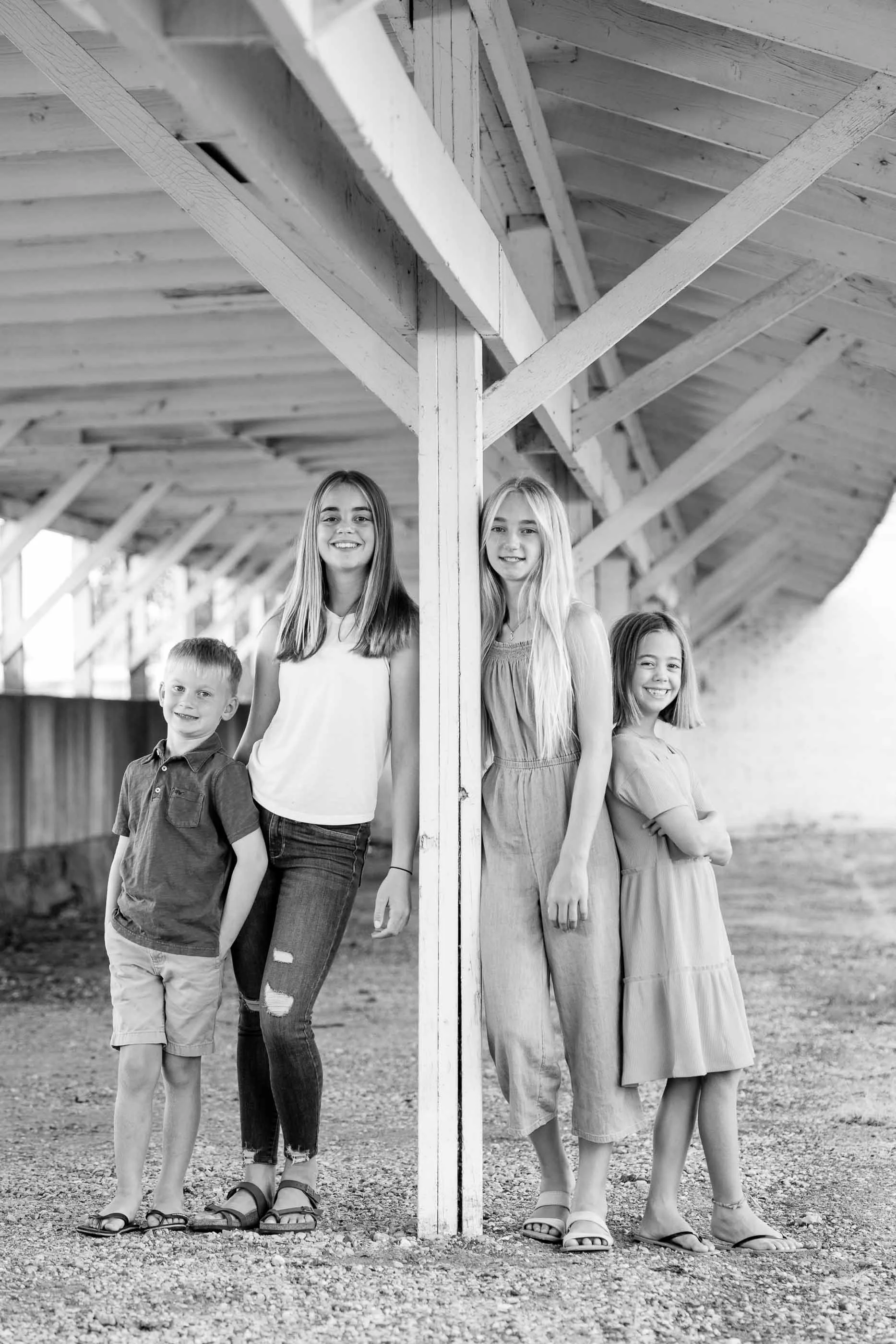 Four children, two boys and two girls, standing by a wooden structure beneath an angled roof, smiling and posing for the photo in black and white.
