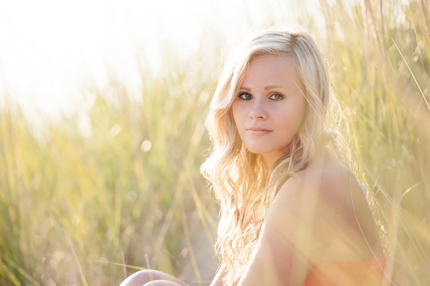 A young woman with long blonde hair sitting outdoors in a field of tall grass, looking at the camera with a soft expression, illuminated by warm sunlight.