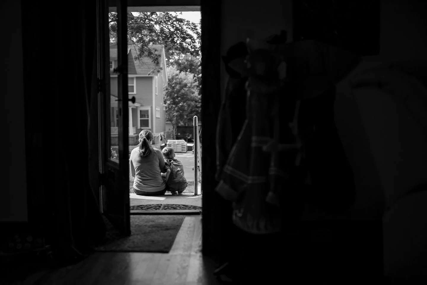 A woman and a young boy sitting on the porch steps outside of a house, looking at the street and neighboring houses.