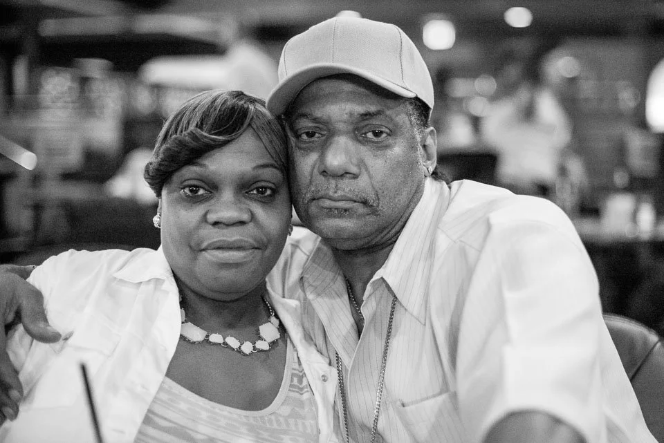 A black and white photo of a man and woman sitting close together, with the man's arm around the woman's shoulder. The woman has short hair and is wearing a necklace and earrings, and the man is wearing a cap and a collared shirt. They are in a setti
