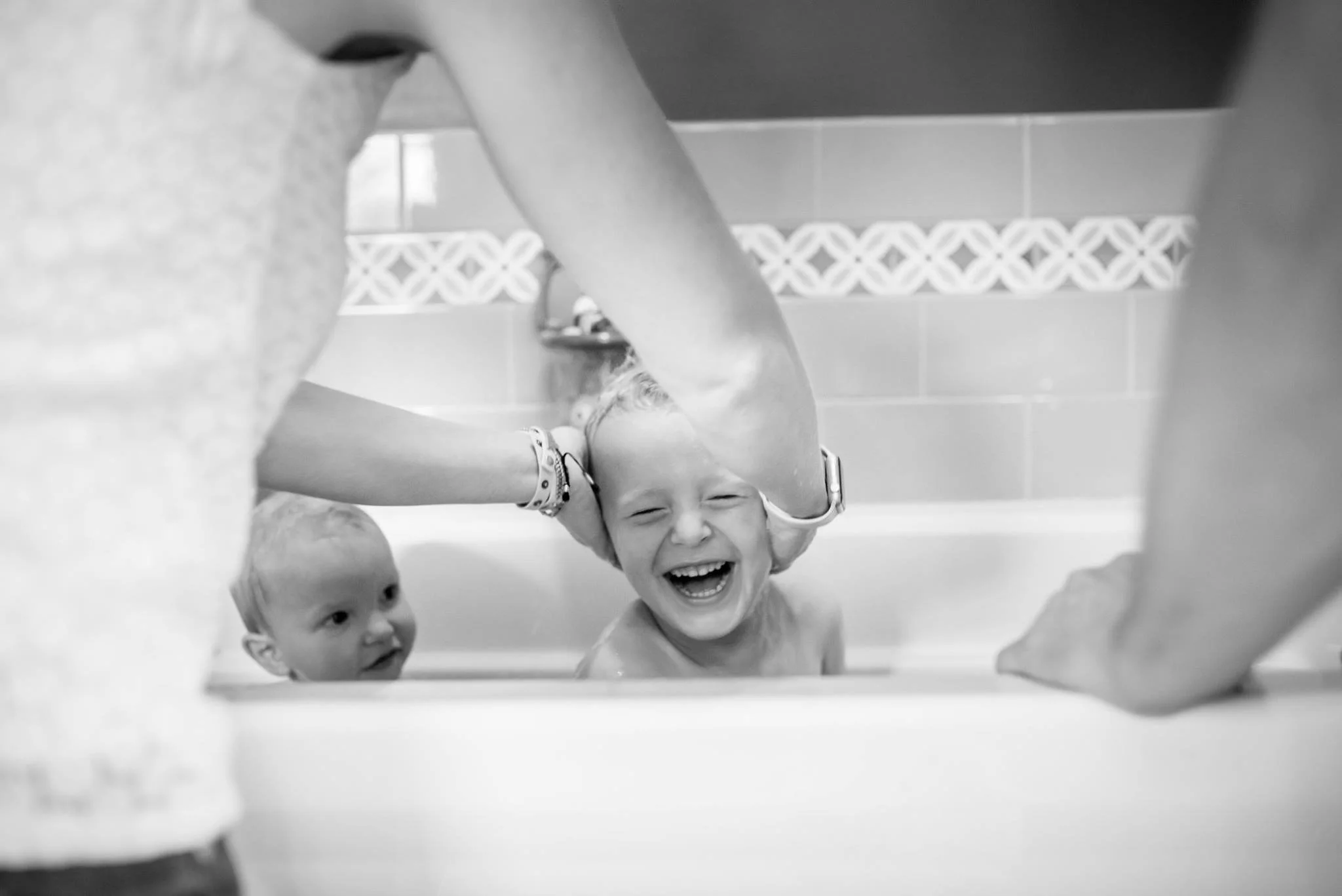 Two children taking a bath, with one child smiling and laughing while being held, and another child looking on.