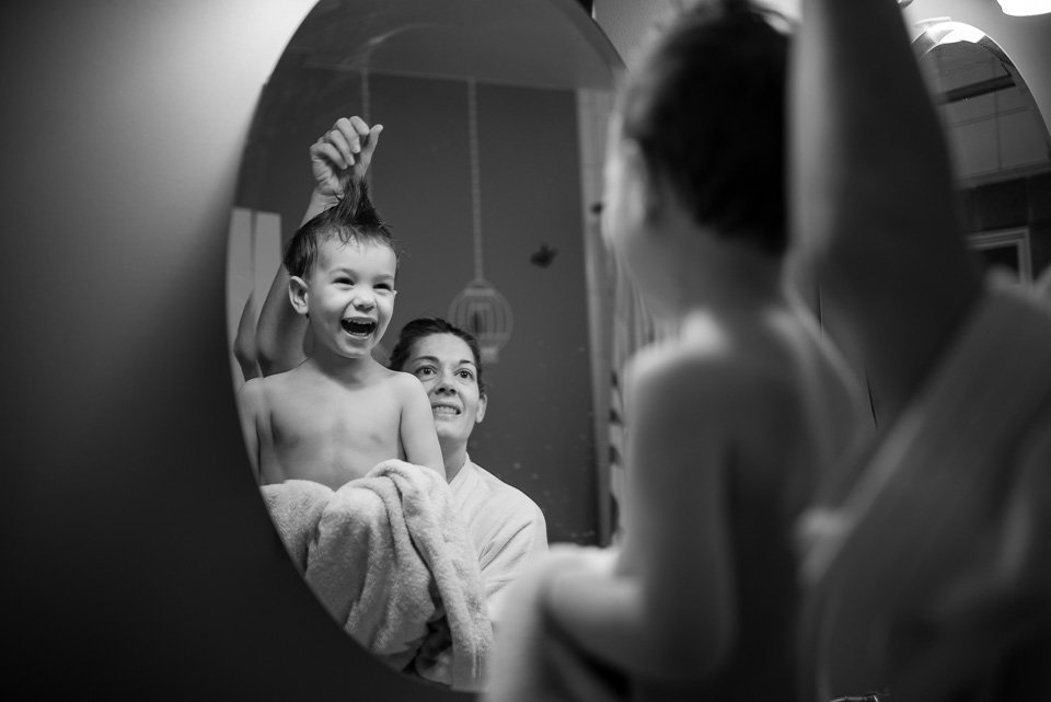 A young boy smiling and laughing while having his hair styled in a salon, with a woman holding his hair up and another woman looking into a mirror in the background.
