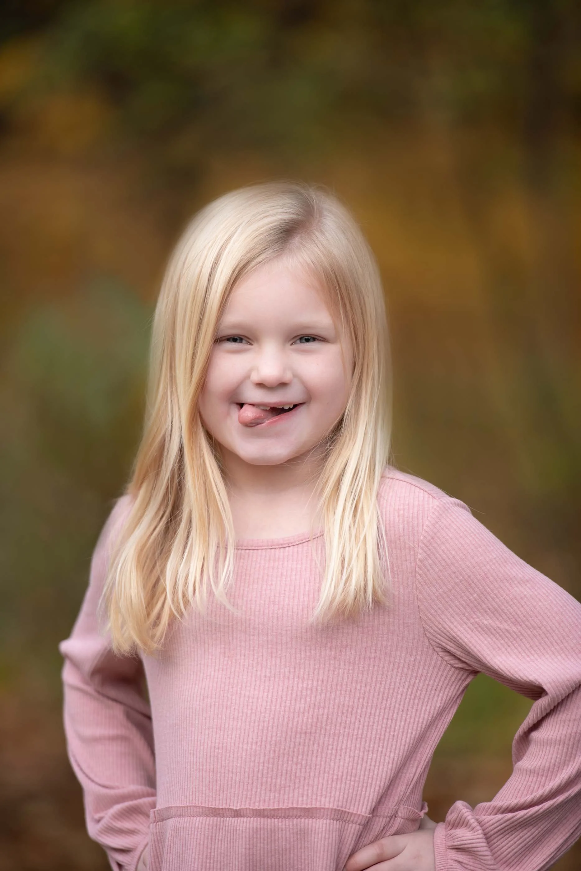 A young girl with long blond hair, wearing a pink long-sleeve shirt, smiling playfully with her tongue slightly out, in an outdoor setting with blurred autumn foliage in the background.