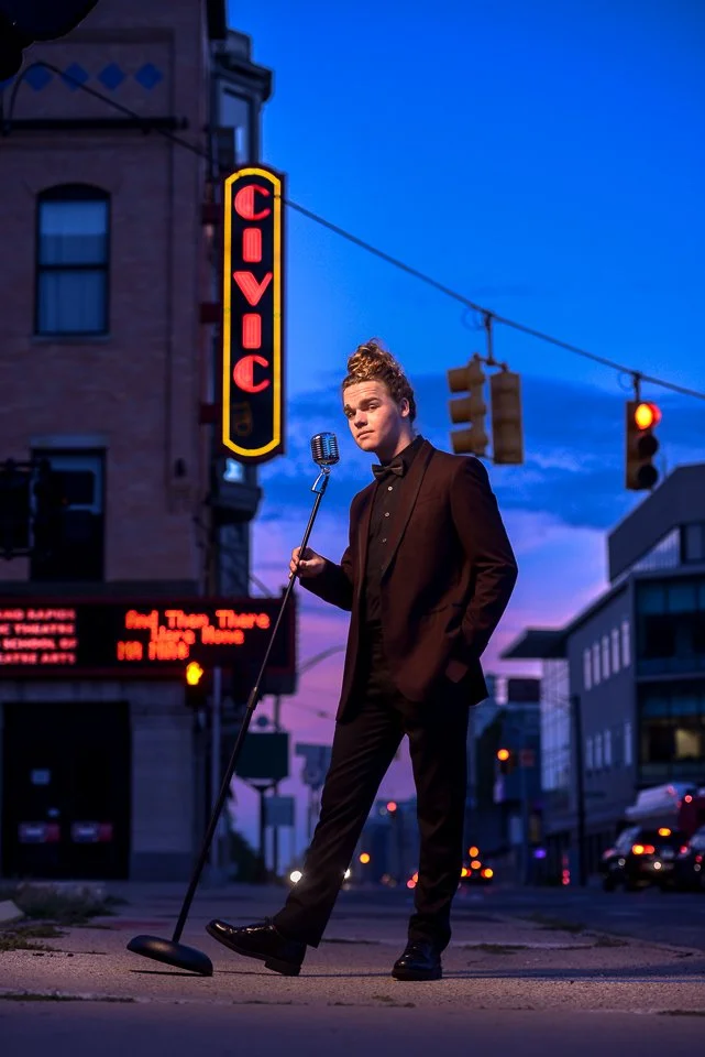 A young man in a black tuxedo with a bow tie stands on a city street at dusk, holding a vintage microphone on a stand. Behind him is an illuminated neon sign that reads "CIVIC" and traffic lights hanging over the street. The sky is a deep blue with f