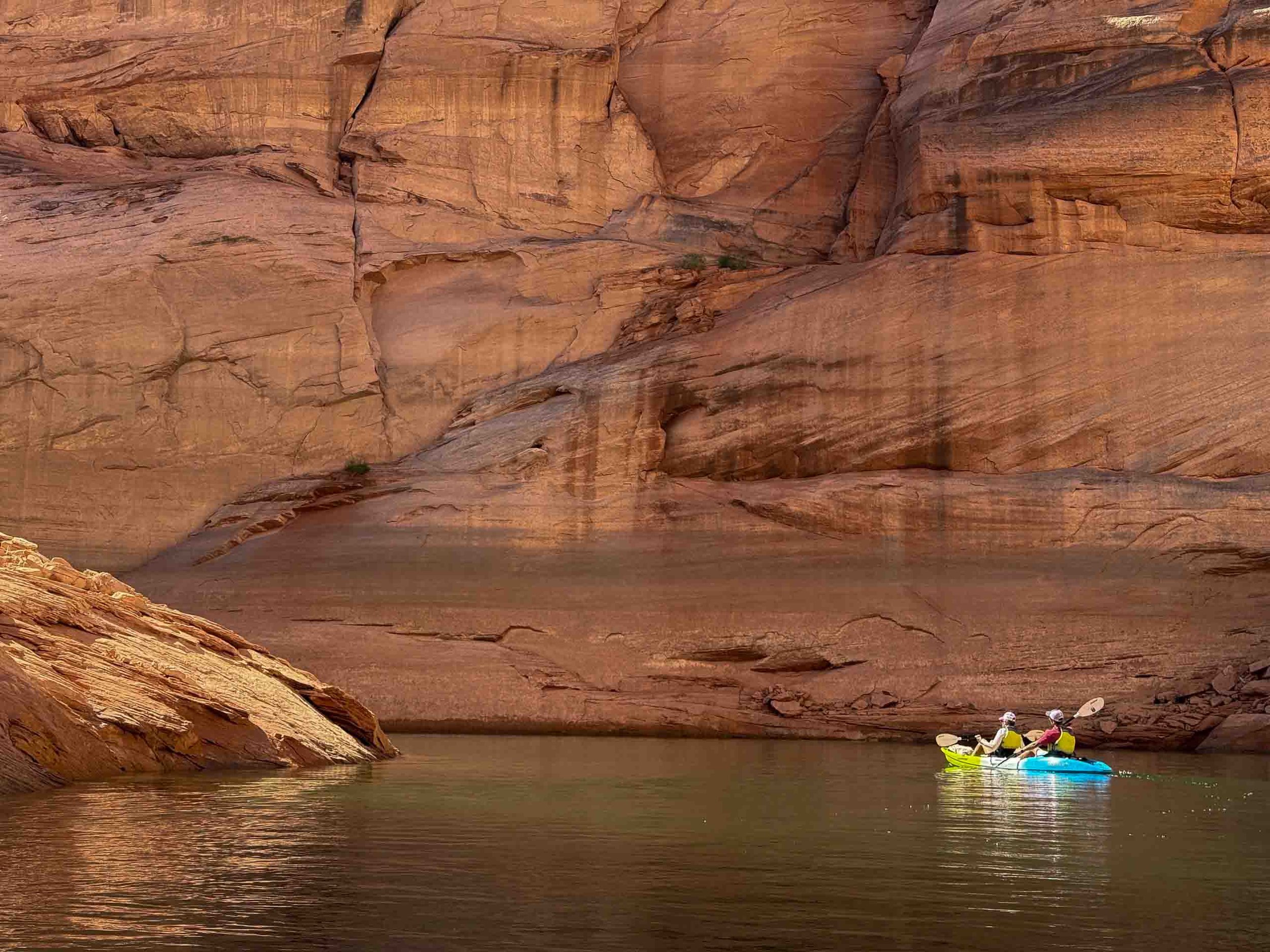 Two people in a yellow and blue kayak paddling on a calm river surrounded by tall red rock canyon walls.