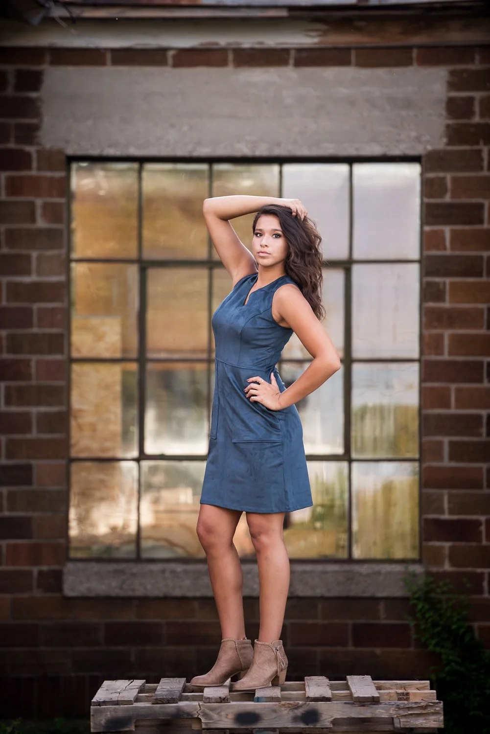 A woman in a sleeveless blue dress and beige ankle boots standing on a wooden pallet outdoors in front of a large window with a brick wall background.