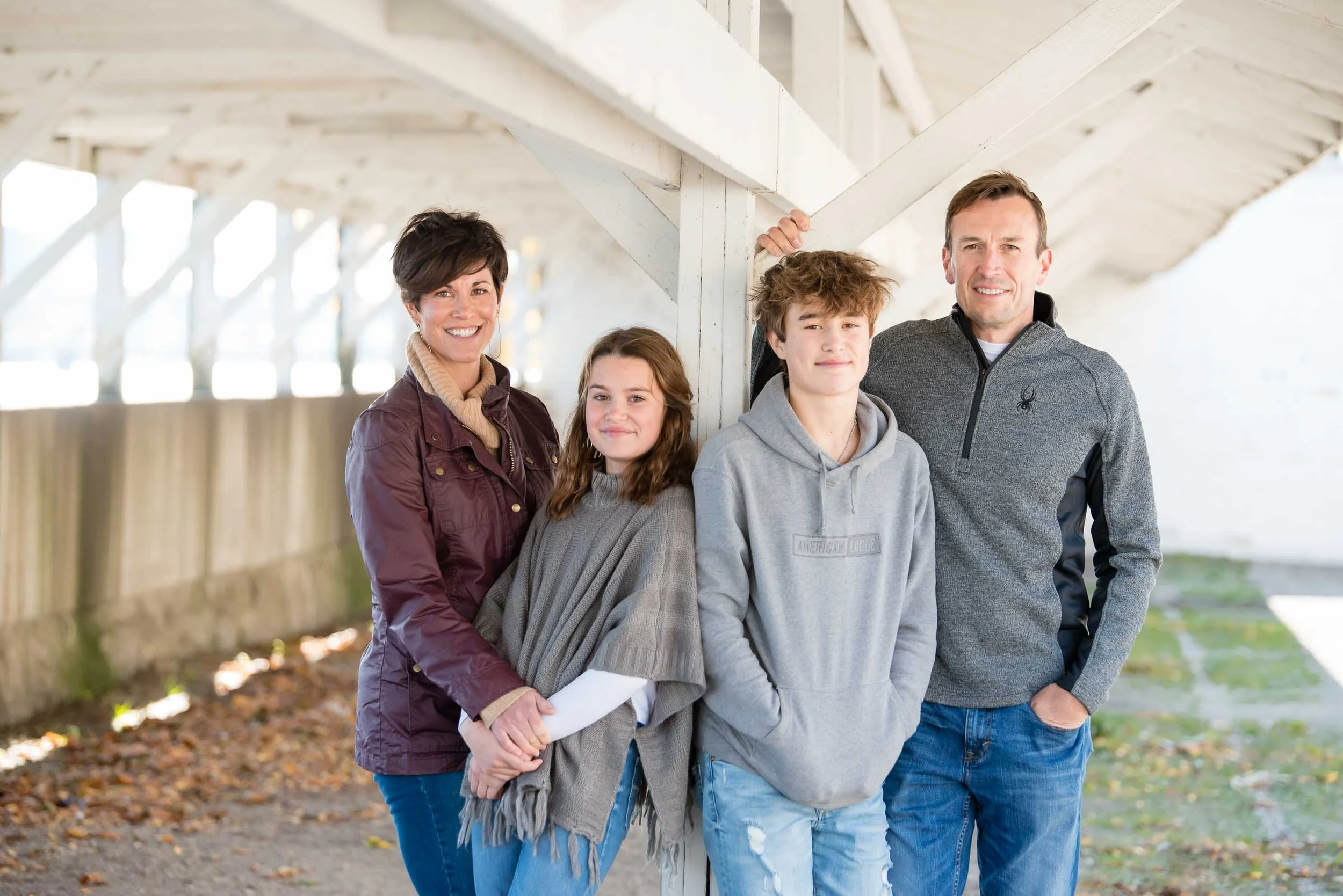 A family of five stands together outdoors under a white wooden bridge, smiling at the camera. The mother is on the left, holding hands with her daughter in the middle. The son and father are on the right, with the father leaning against the bridge su