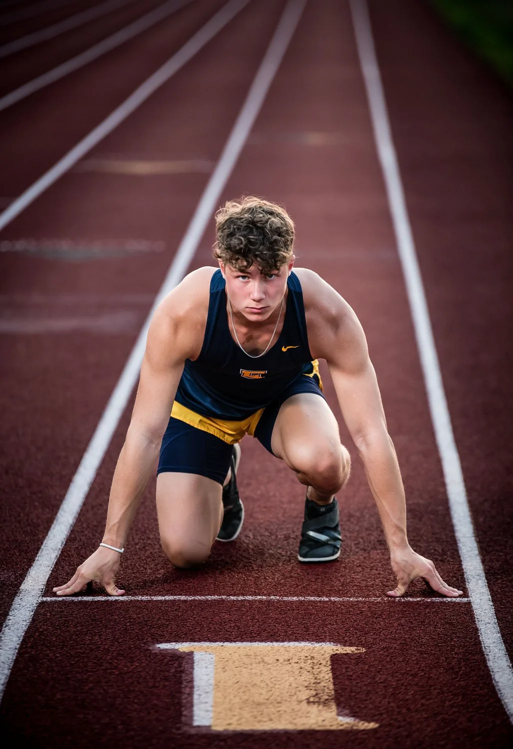 A young male athlete in navy and yellow sportswear crouches on a red running track, ready to start a race, with white lane markings and a serious expression.