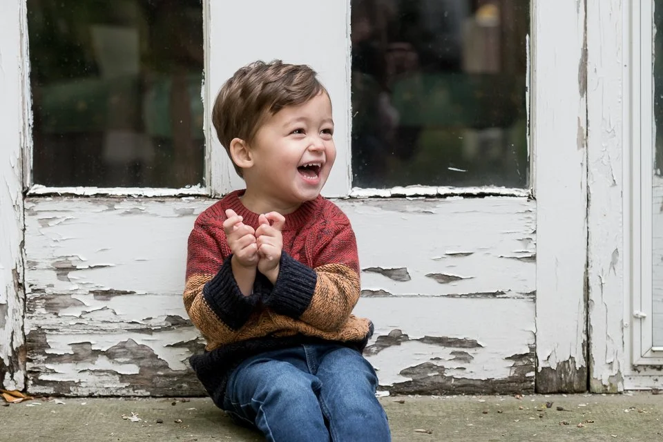 A young boy sitting on a porch, laughing happily, with peeling white paint on the door behind him.