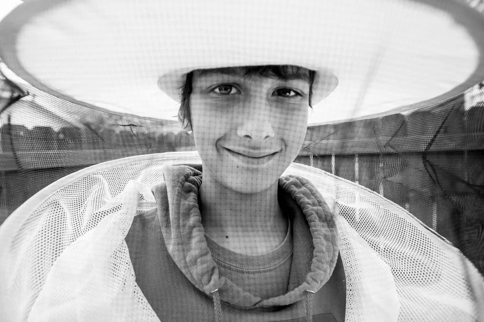 Young person smiling, wearing a wide-brimmed hat and hoodie, outdoors with a netted structure nearby.
