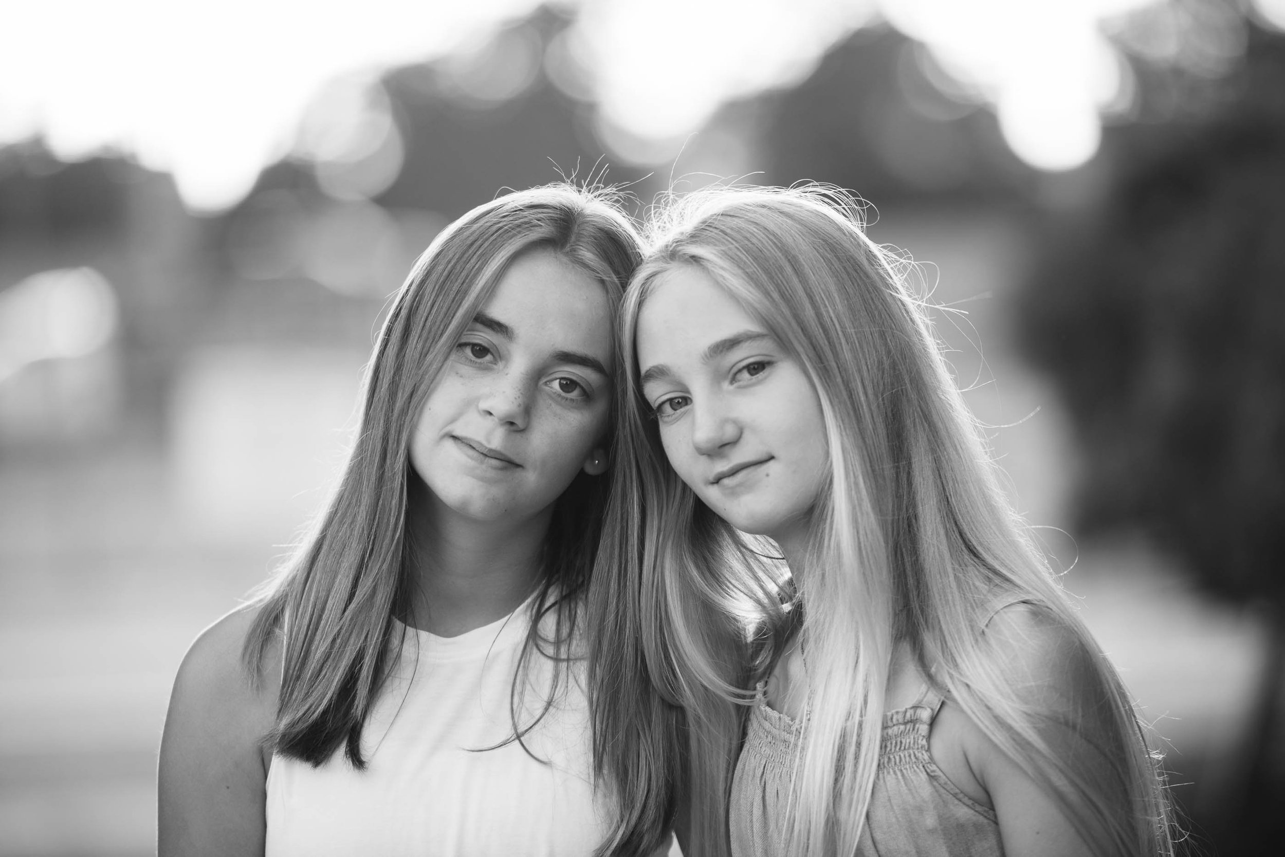 Black and white photo of two young girls with long hair, smiling gently, standing close together outdoors with blurred trees and sky in the background.