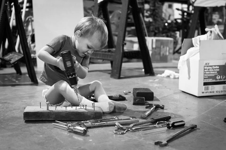 A young boy sitting on the floor using a power drill with various tools scattered around him in a garage or workshop.