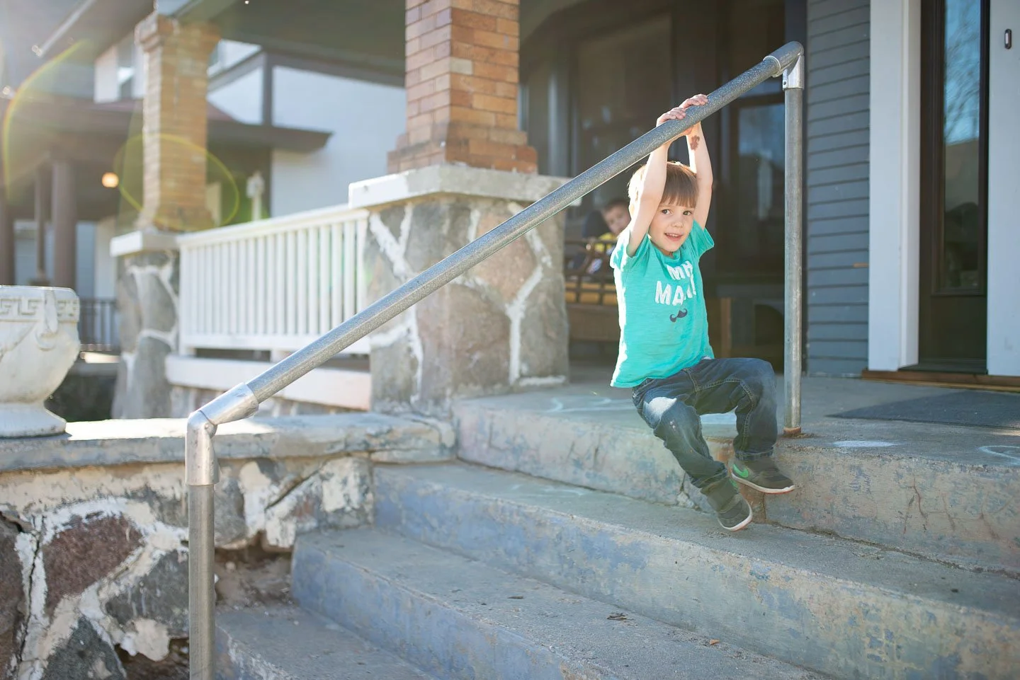 A young boy sitting on a concrete staircase near a house, holding onto a metal handrail, with a bright smile on his face.