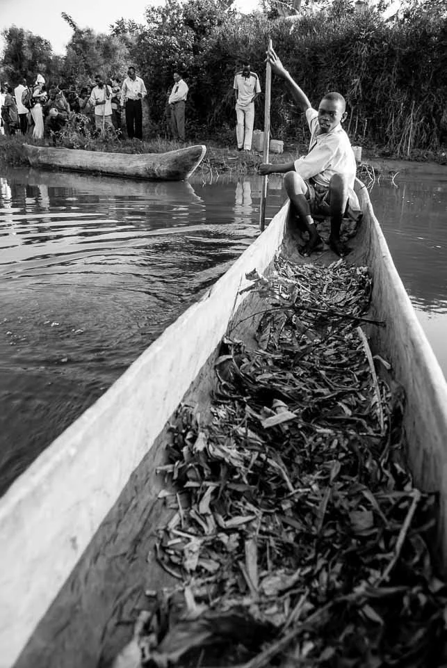 A young boy sitting in a narrow wooden boat filled with leaves and debris, holding a paddle with one hand and raising the other hand in a peace sign, on a river. Several adults stand on the riverbank in the background.