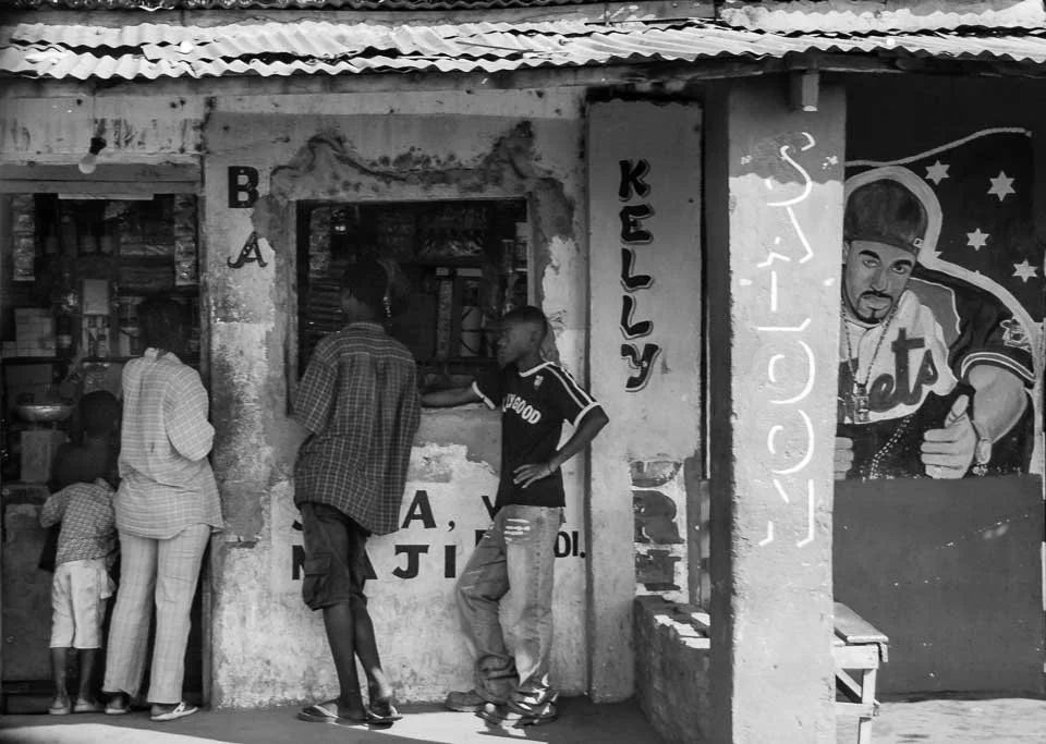 Black and white photo of a storefront with people gathered outside. The building has signs reading 'KELLY' and 'ECLECTICS' and a mural of a rapper with a cap and chain.