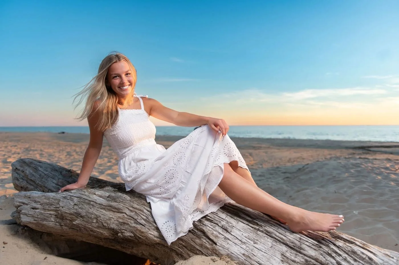 Senior girl in a white dress sitting on driftwood on a Lake Michigan beach at sunset during her senior portrait session