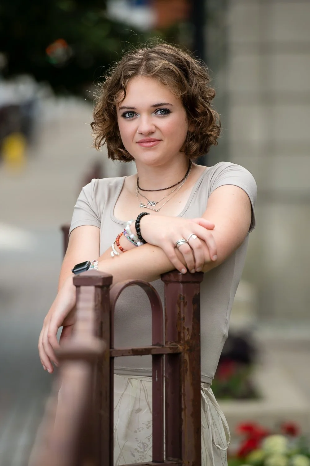 A young woman with curly brown hair and blue eyes leaning on a rust-colored metal railing outdoors.