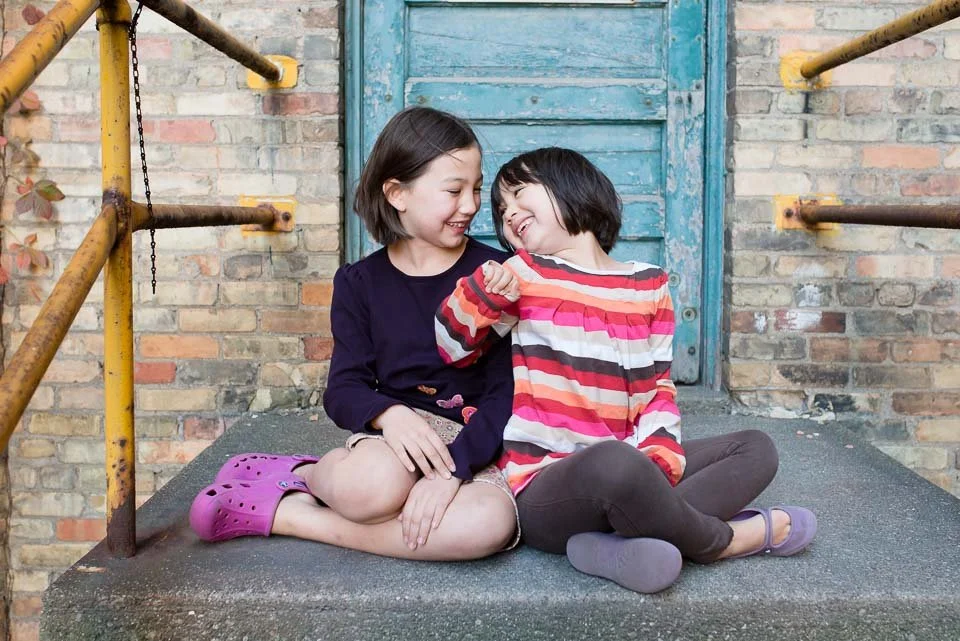 Two young girls sitting on a concrete step in front of a brick wall with a weathered blue door, smiling and playing together