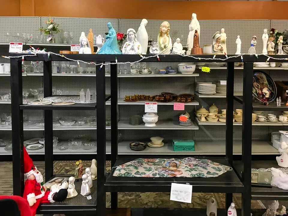 Various Christmas and religious-themed figurines and ceramics on display in a thrift store, with shelves filled with dishes and decorative items in the background.