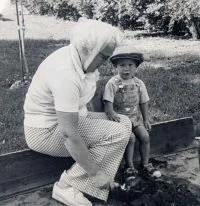 An elderly woman with glasses and short hair is sitting on a bench outside, talking to a young boy wearing a hat, both looking at each other. There are trees and a grassy area in the background.