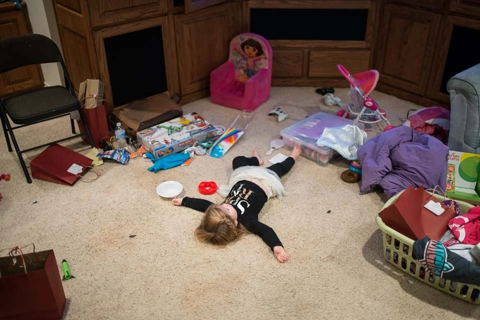 Child lying on the carpet with scattered toys, several bags, and a pink doll chair in a cluttered room.