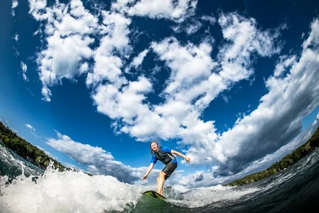 Person surfing on a wave in a river under a partly cloudy sky.