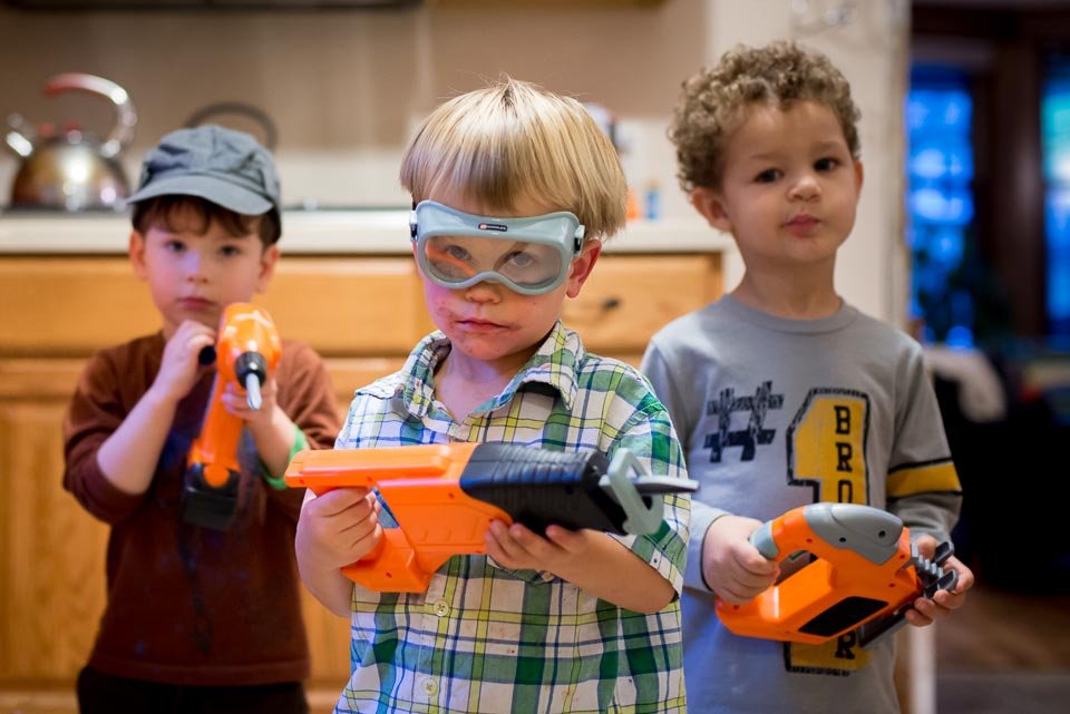 Three young boys playing with toy guns indoors, one boy wearing goggles and a plaid shirt, the second boy in a gray shirt, and the third in a brown shirt and cap.
