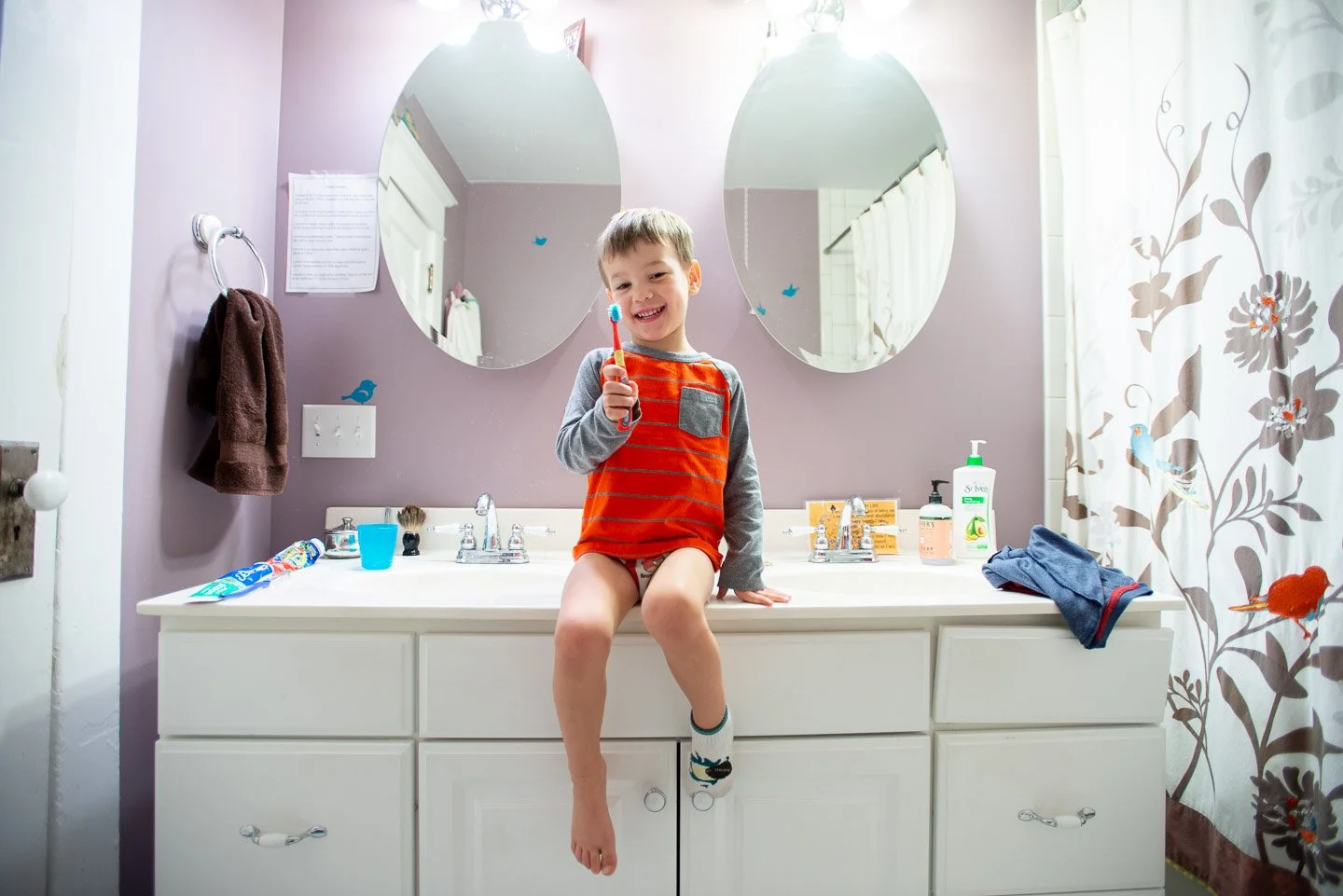 A young boy sitting on a bathroom sink countertop, holding a toothbrush and smiling. The bathroom has two mirrors, a purple wall, and a curtain with bird and floral patterns.