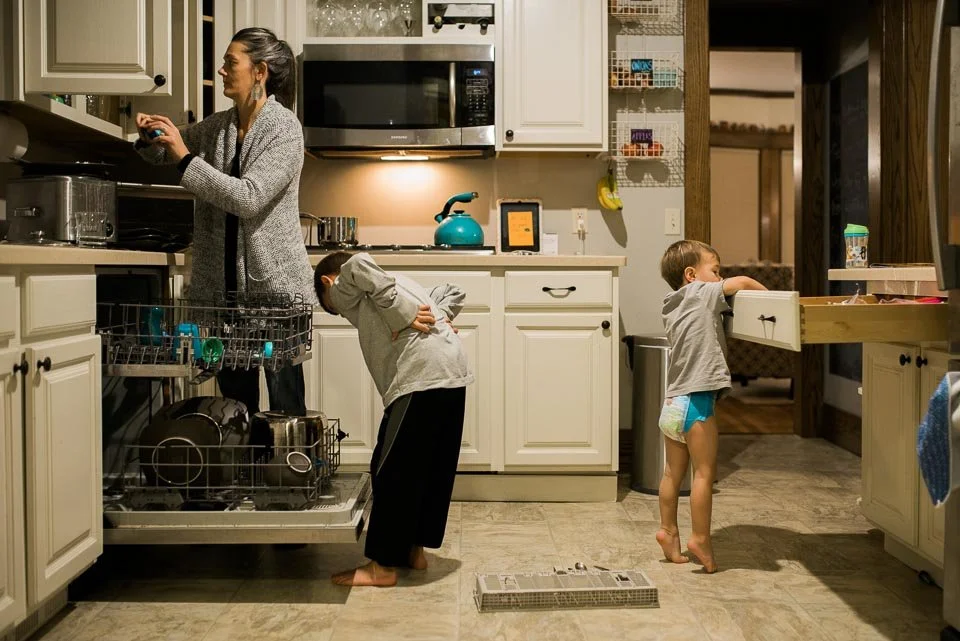 A woman is preparing food in the kitchen while two children are distracting her, one leaning into a drawer and the other standing with arms on the counter.