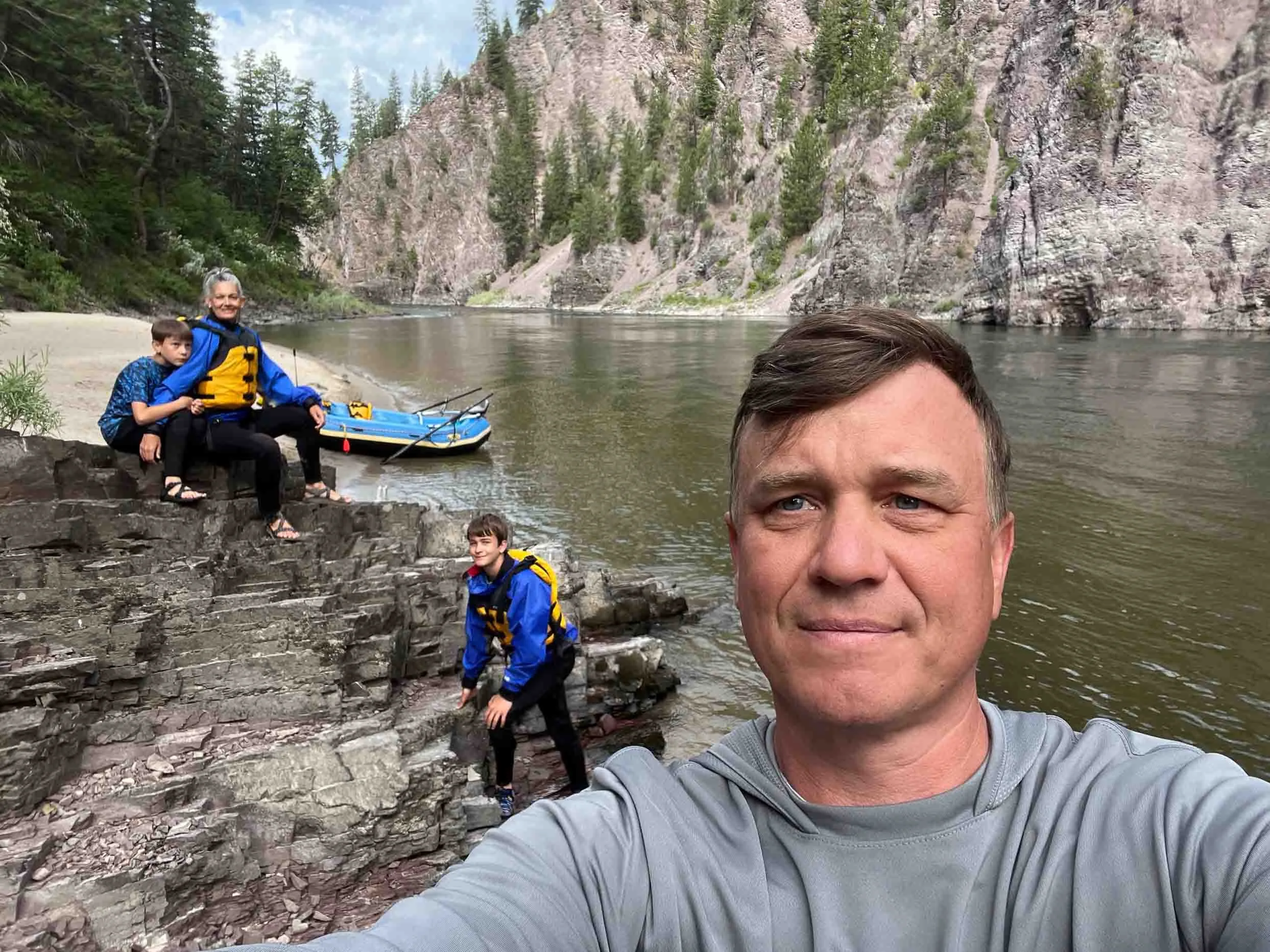 A man taking a selfie outdoors near a river with three kids and a canoe, surrounded by trees and mountains.