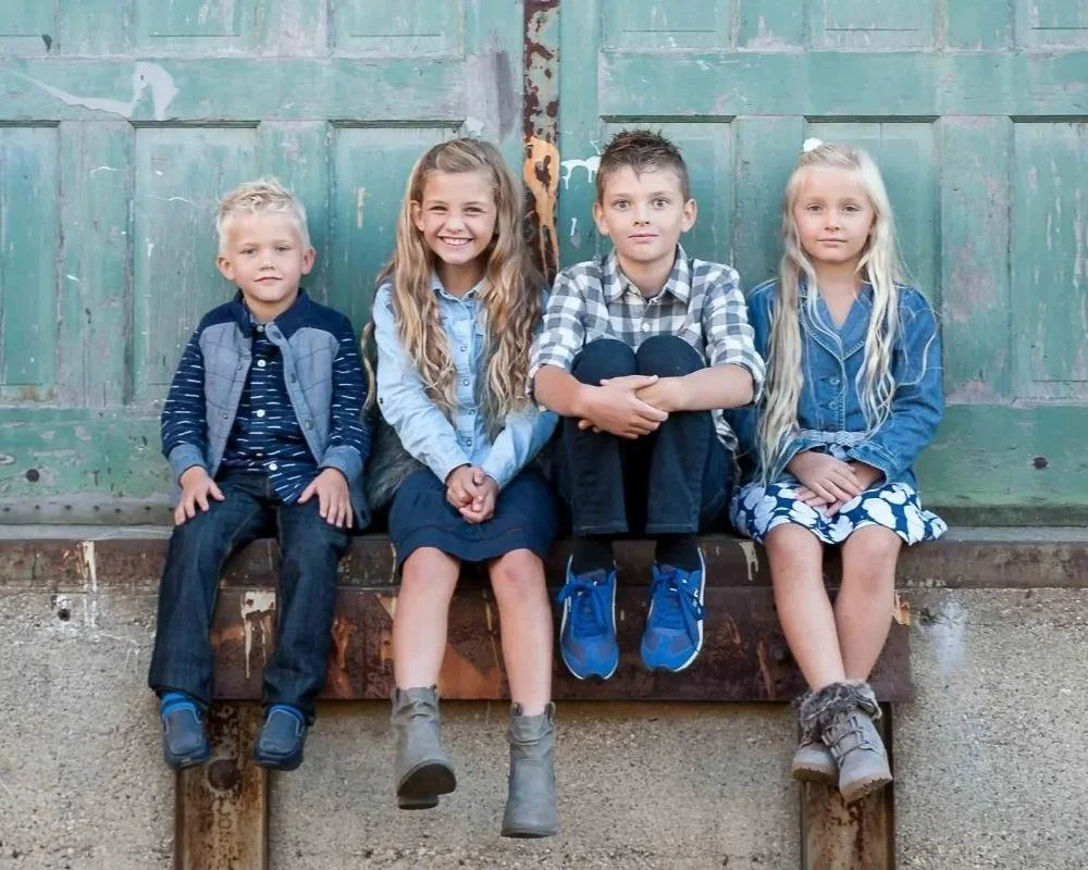 Grooters family of four children sitting together on a rustic bench against weathered green doors
