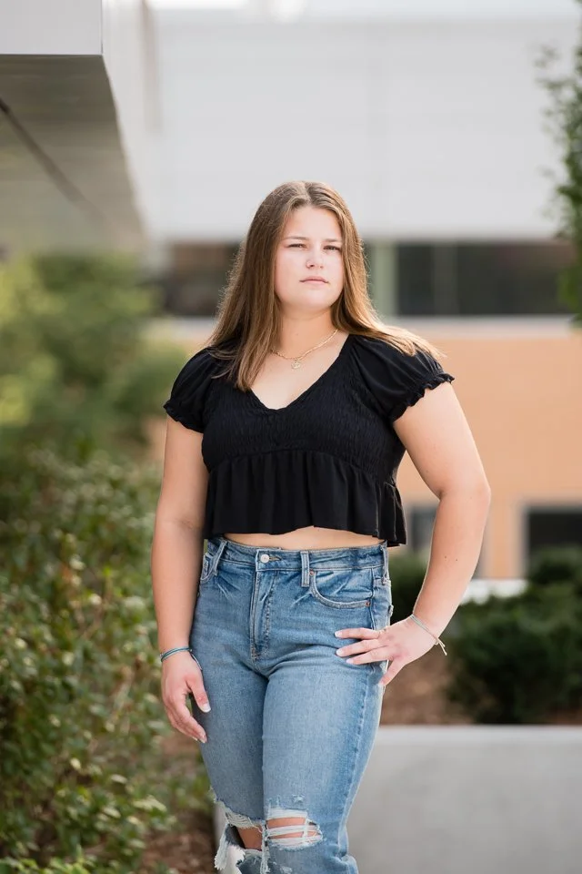 A young woman with long brown hair standing outdoors in a casual outfit, wearing a black crop top and ripped jeans, with one hand on her hip and a neutral expression.
