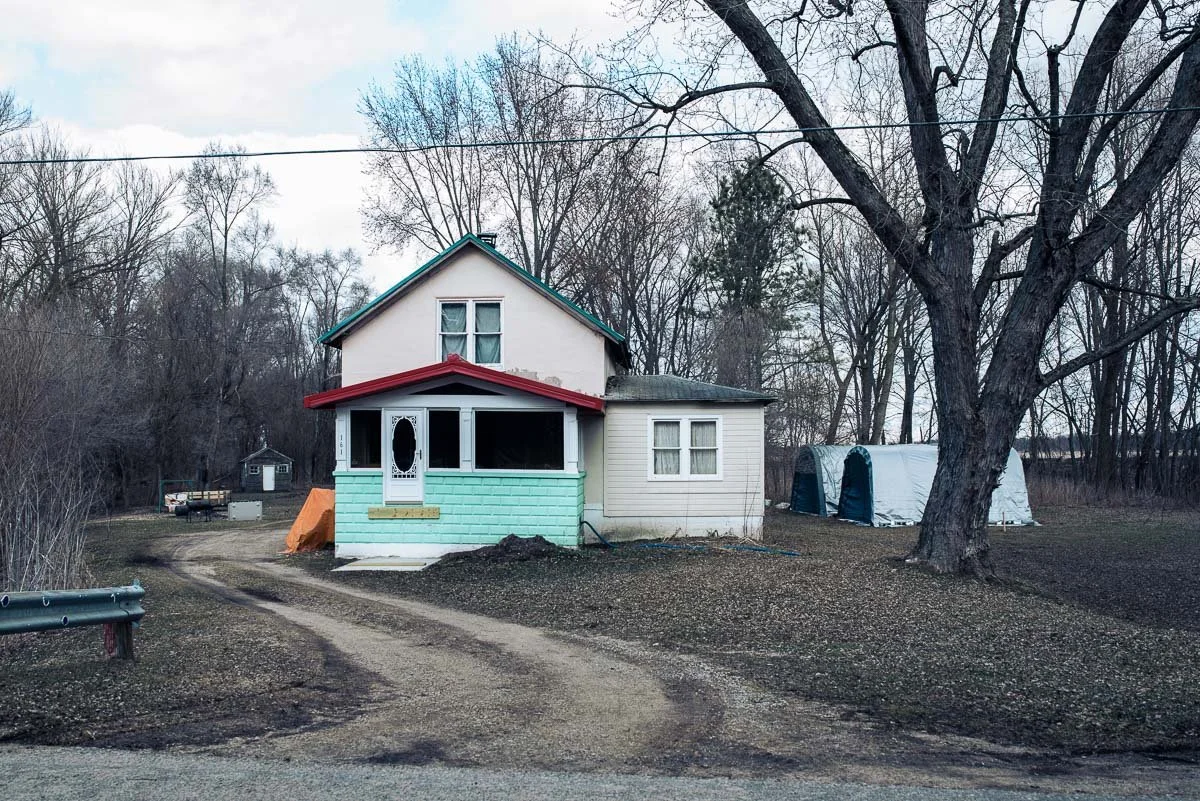 A small two-story house with a covered porch surrounded by leafless trees and a dirt driveway, with a large tree in the yard and two small tents or structures in the background.