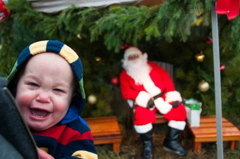A crying toddler in colorful hoodie in front of Santa Claus sitting on a bench under a decorated Christmas tree.