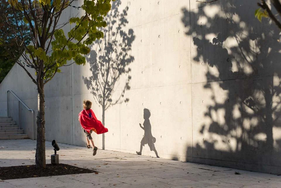 A young child wearing a red cape is running on a paved sidewalk, casting a shadow that looks like they have a cape fluttering behind them. There are trees and stairs nearby, with their shadows cast on a concrete wall.