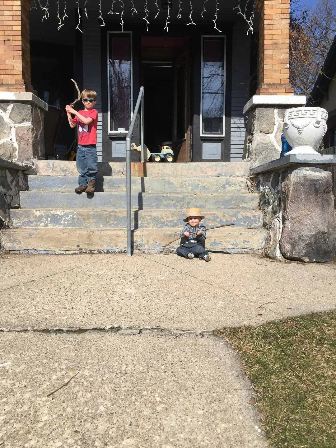 Two young children on the front steps of a house, one standing at the top of the stairs in a red shirt and sunglasses holding a stick, the other sitting on the ground in a hat and striped shirt holding a stick, with toys and decorations visible in th