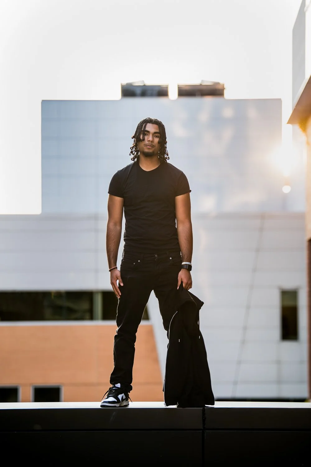 Young man with black shirt and pants, holding a jacket, standing on a ledge outdoors in front of modern buildings during sunset.
