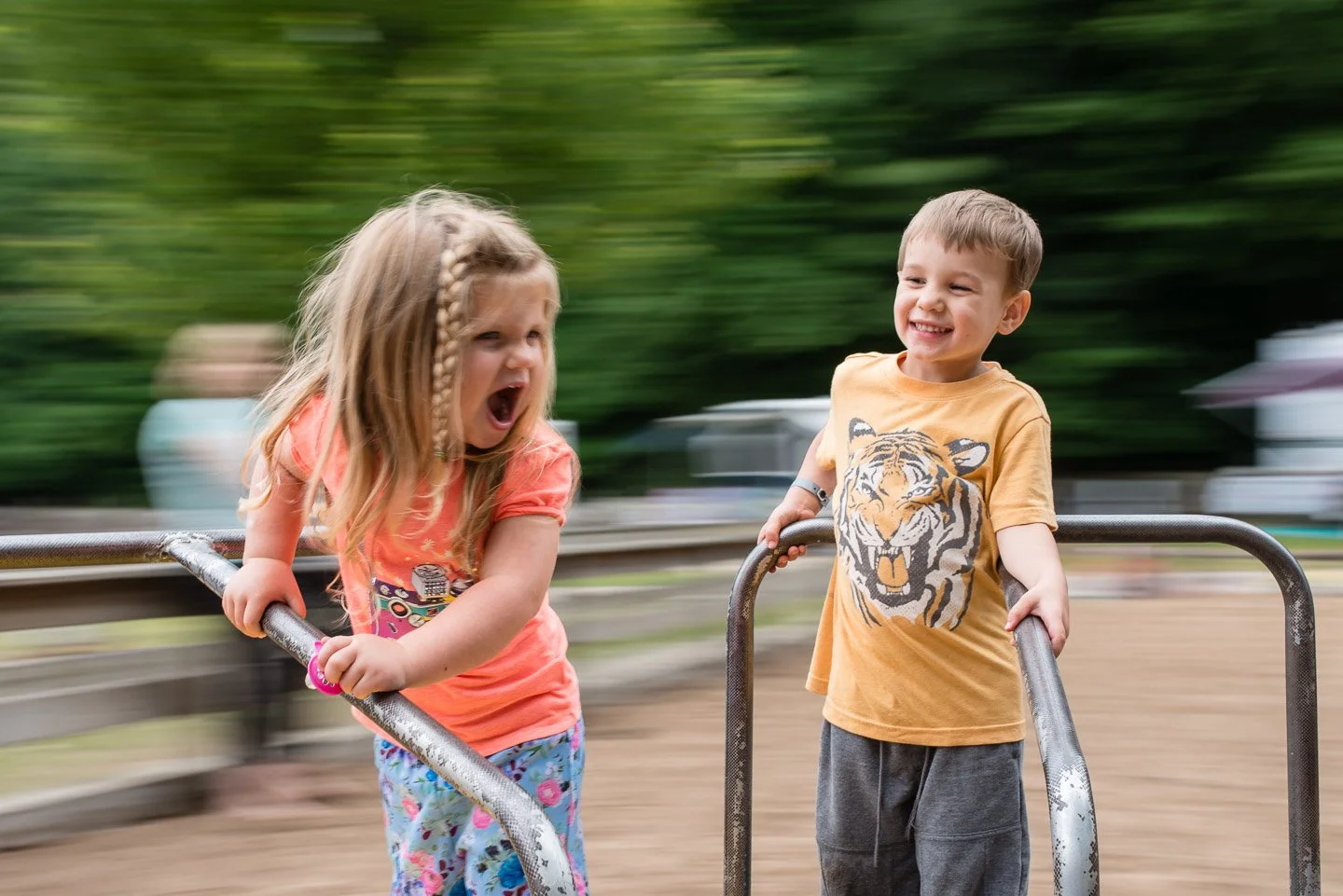 Two children, a girl and a boy, are playfully pushing each other on a metal merry-go-round at a park, with blurred trees and other park features in the background.