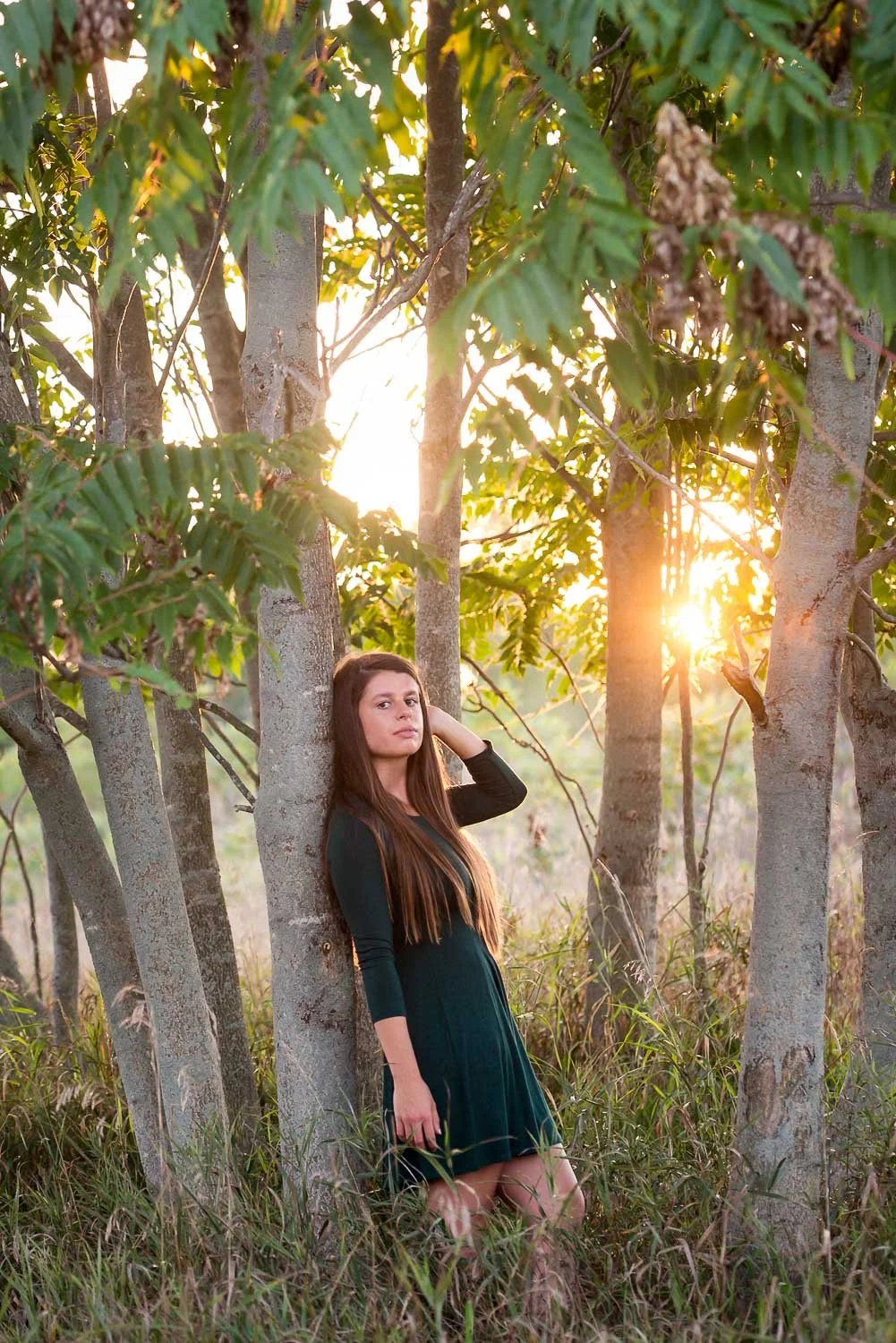 A woman standing among trees in a grassy area at sunset, wearing a dark green dress, with brown hair, looking at the camera.