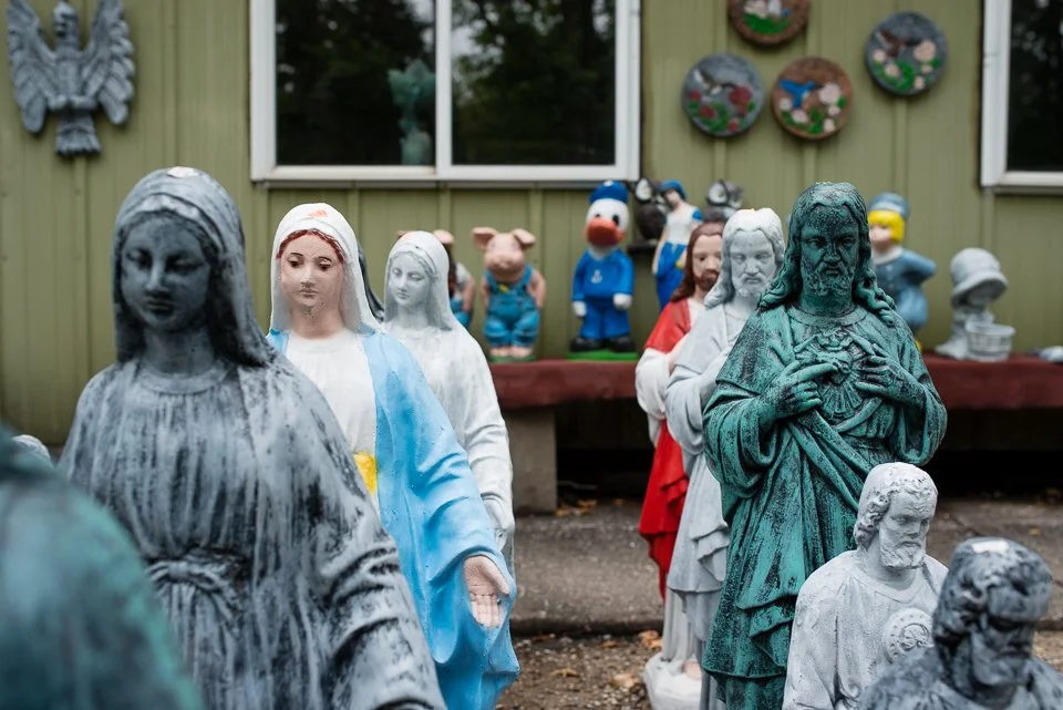 Collection of religious and cartoon character statues displayed outdoors against a green wooden building with window and decorative plates and a shelf.