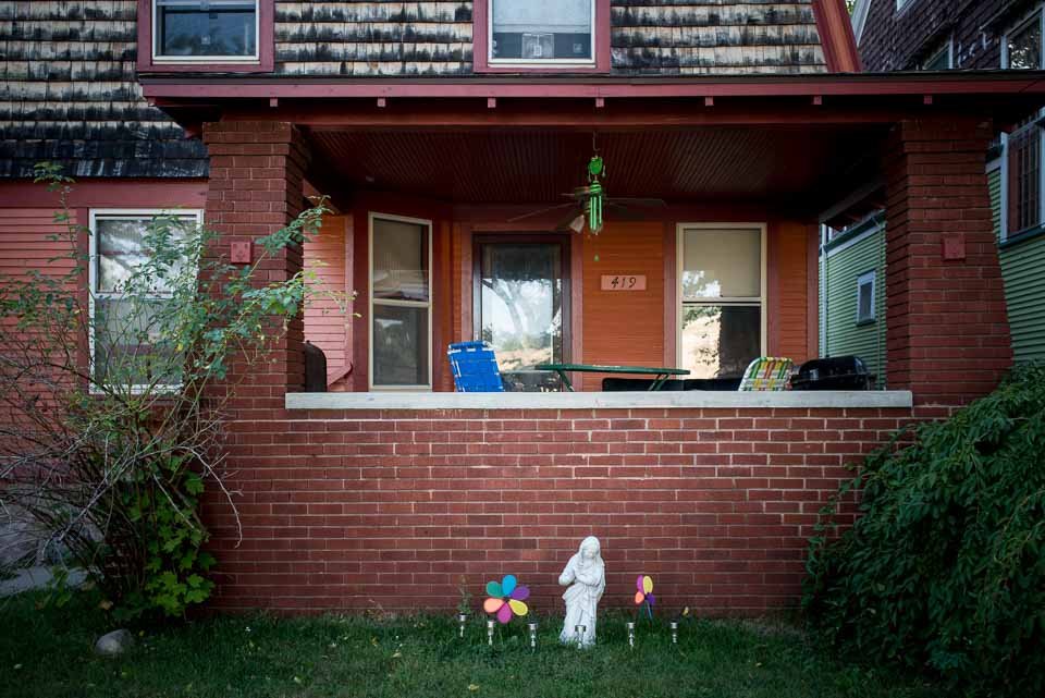 Front porch of a brick house with a covered balcony. There are two chairs, a table, and a grill on the porch. In front, on the lawn, there are four colorful pinwheels and a small statue of a woman.