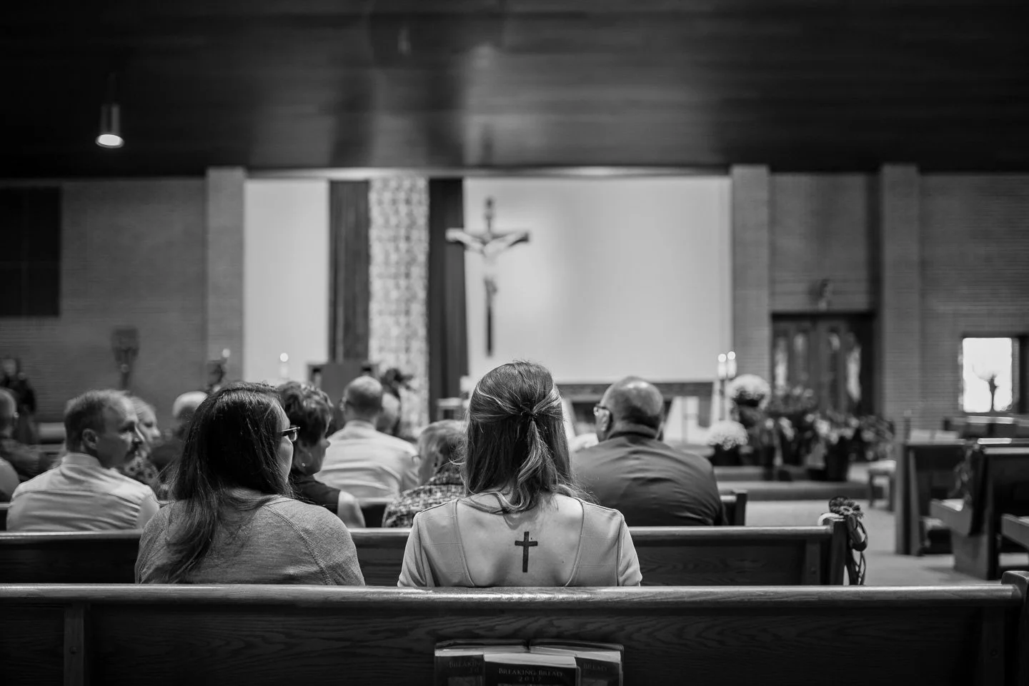 People sitting in pews inside a church, attending a service with a crucifix on the wall in the background.