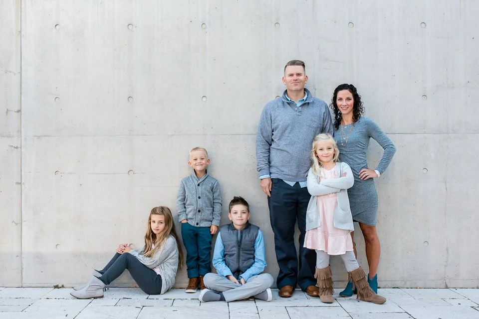 Family of six standing and sitting against a concrete wall, smiling, with three children sitting on the ground and two adults standing behind them.