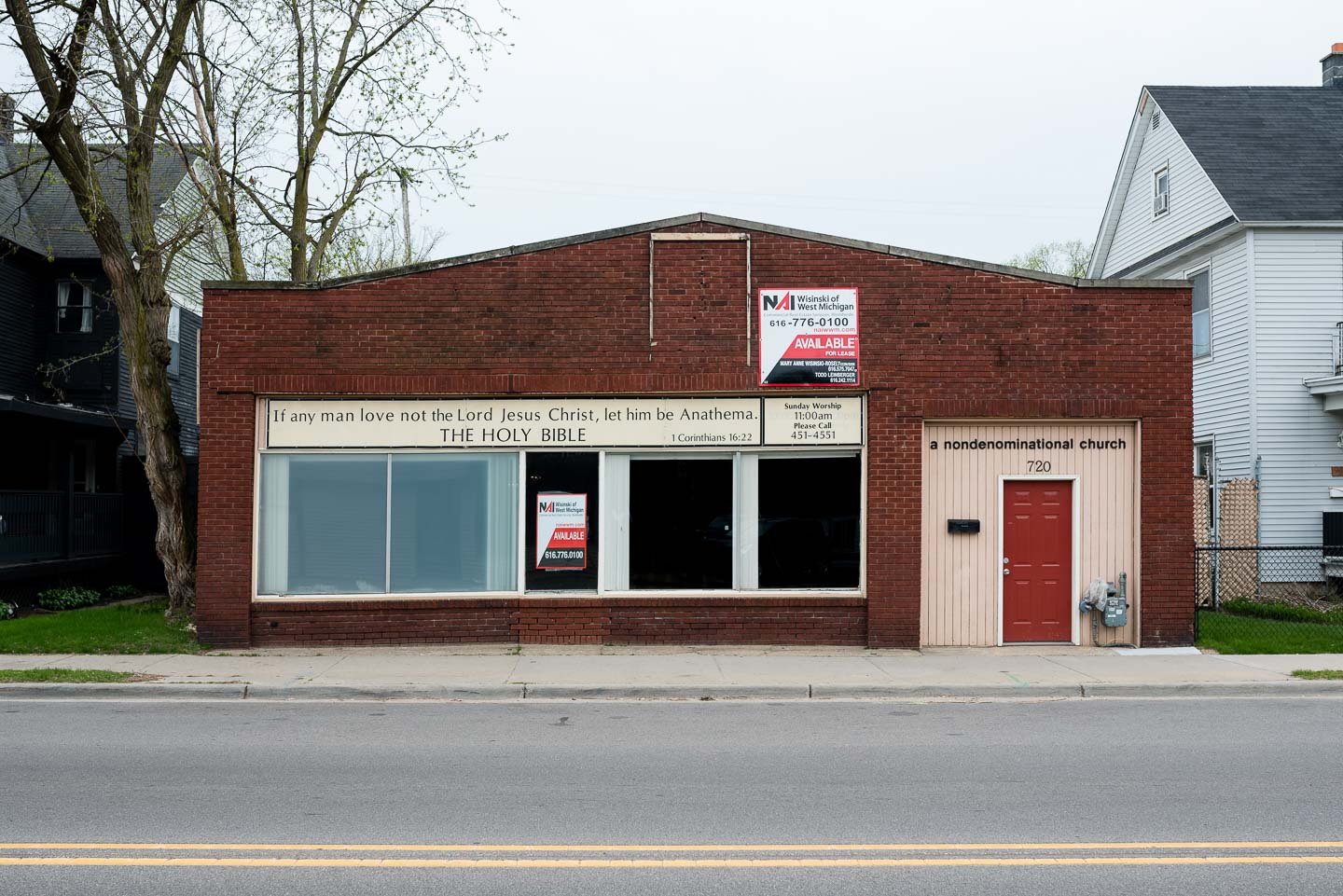 The exterior of an unassuming single-story brick church with a pinkish door, signage indicating it's a non-denominational church, and a large window with signs. The church's address is 720. A biblical quote from 1 Corinthians 16:22 and worship time a